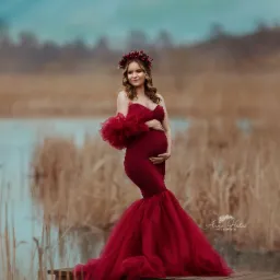 Kobieta w ciąży w czerwonej sukni z falbanami, stojąca na drewnianym pomoście nad wodą, w tle trzciny i pochmurne niebo, na głowie wianek z czerwonych kwiatów.