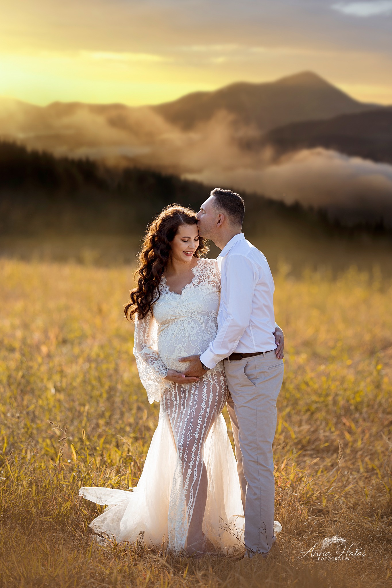 Przyszła mama w koronkowej sukni i tata w jasnych spodniach na tle gór o zachodzie słońca, mężczyzna całuje kobietę w czoło.
