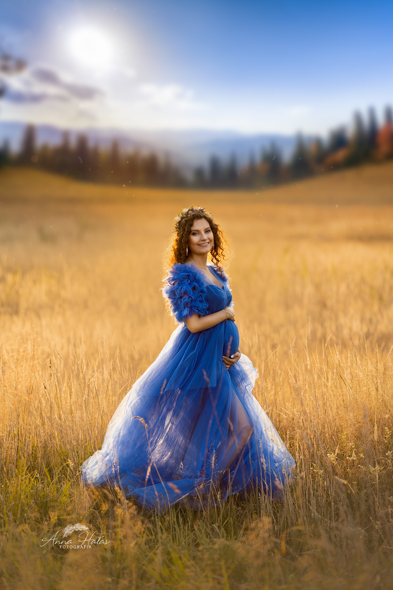 Uśmiechnięta kobieta w ciąży, w błękitnej, zwiewnej sukni, stoi na polu w złocistym świetle zachodzącego słońca, z wiankiem na głowie i rękami na brzuchu.
