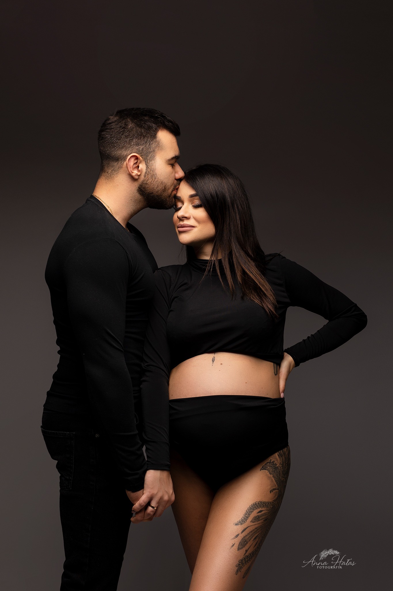 Mężczyzna w czarnej koszulce całuje w czoło kobietę w czarnym topie, widoczny ciążowy brzuszek i tatuaż feniksa na udzie, szare tło.