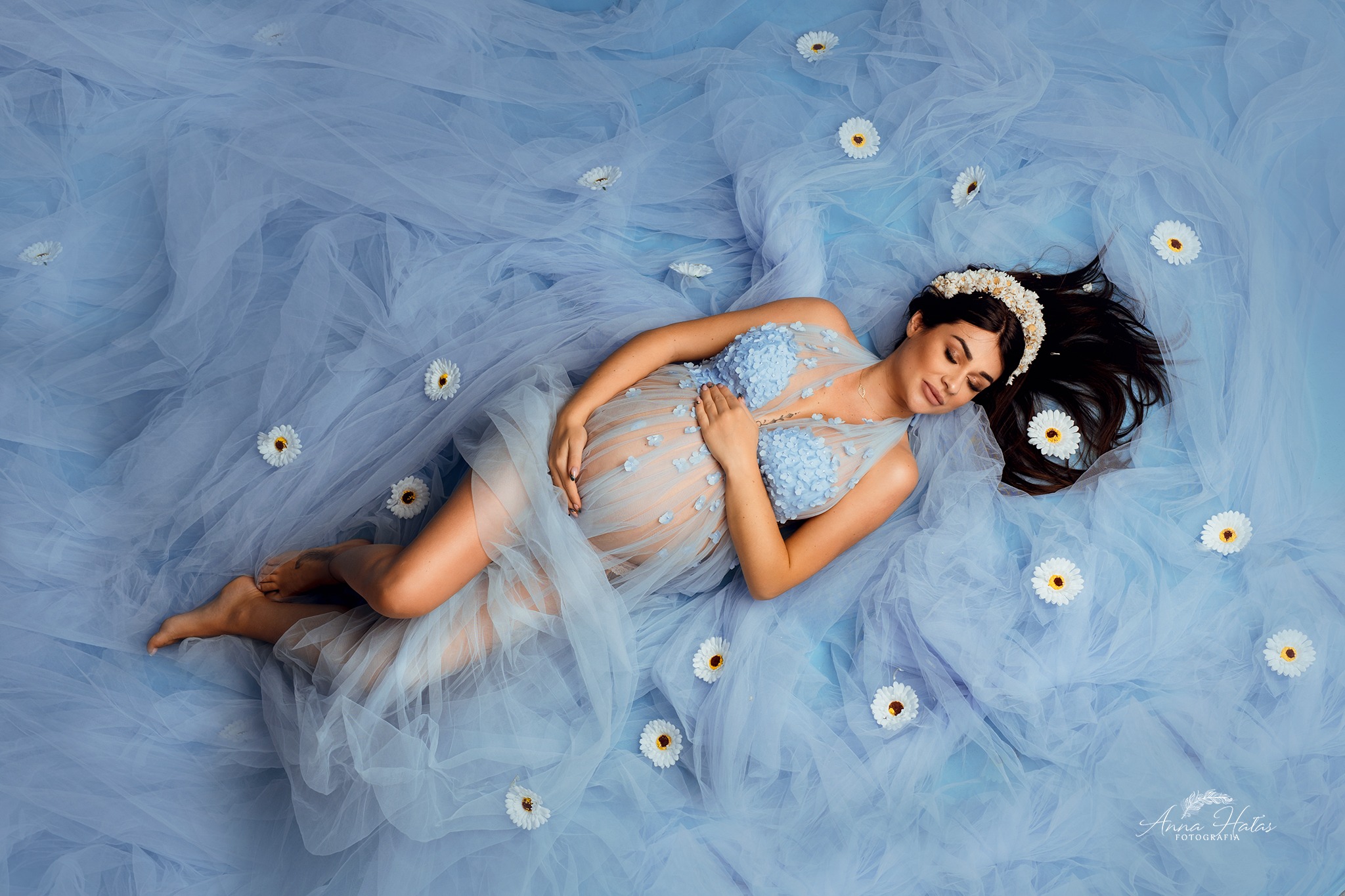 Kobieta w ciąży leży na błękitnym tiulu z rozrzuconymi stokrotkami, ubrana w delikatną sukienkę z aplikacjami kwiatowymi i wianek na włosach, dłonie spoczywają na brzuchu.