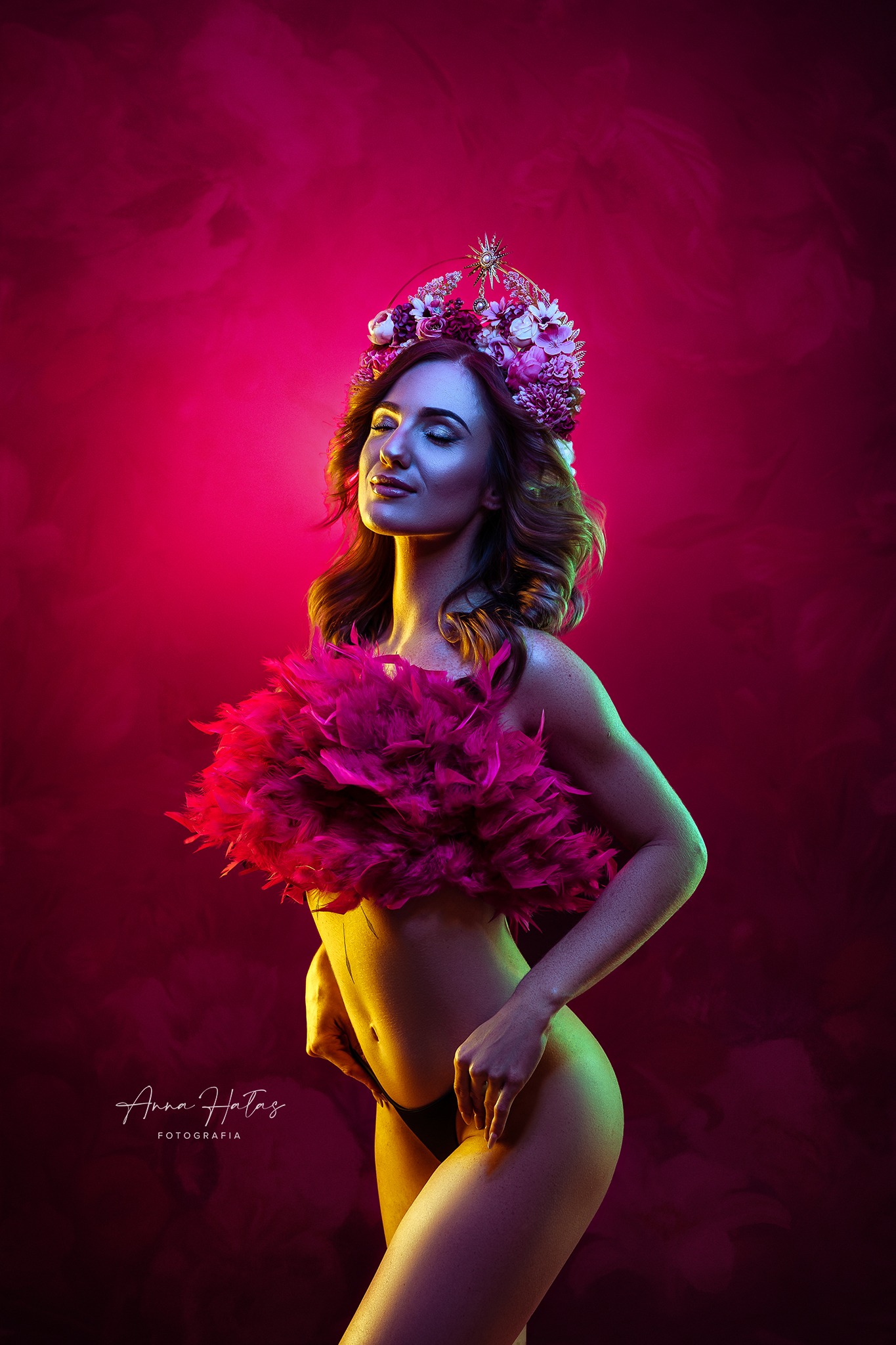 Kobieta z zamkniętymi oczami w koronie z kwiatów i piórach na tle różowego dymu, oświetlona żółto-zielonym światłem.