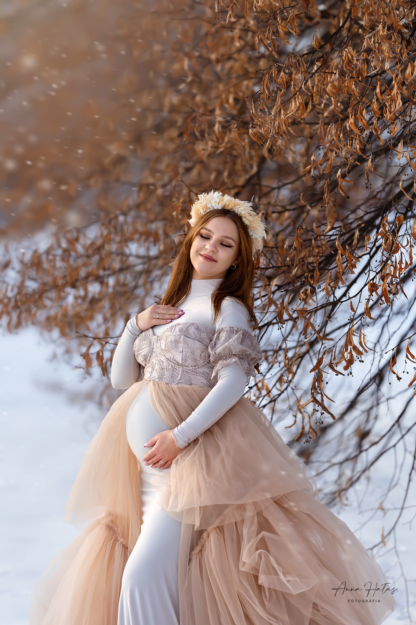 Uśmiechnięta kobieta w ciąży w beżowej sukni z tiulu i wianku na głowie, pozuje na śniegu pod drzewem z brązowymi liśćmi podczas delikatnych opadów śniegu.