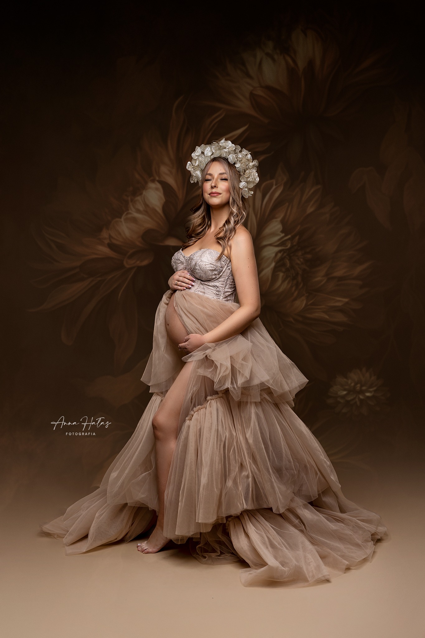 Kobieta w ciąży pozuje w długiej, warstwowej sukni z tiulu w kolorze beżowym i wianku z białych kwiatów na głowie na tle brązowej ściany z motywem kwiatowym.