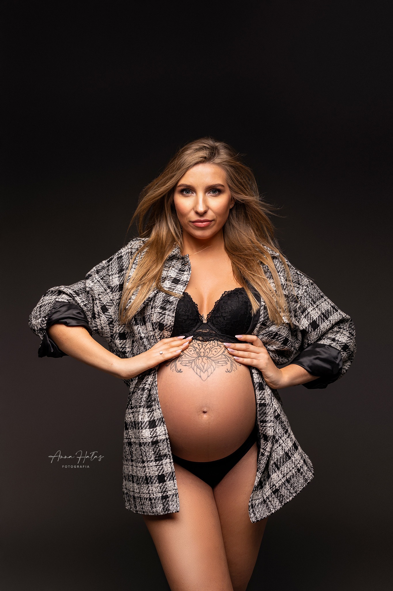 Portret kobiety w ciąży, ubrana w rozpiętą koszulę w kratę i czarną bieliznę, z tatuażem na brzuchu, pozująca na ciemnoszarym tle.