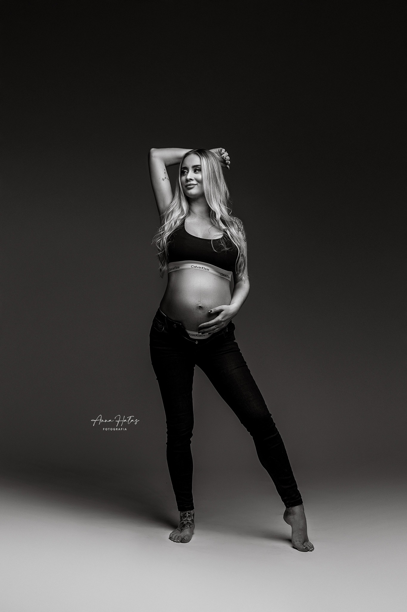 Czarno-białe zdjęcie kobiety w ciąży, ubrana w sportowy stanik i dżinsy, pozująca z jedną ręką uniesioną nad głową, ukazująca tatuaż na ramieniu oraz ozdobny tatuaż na stopie.