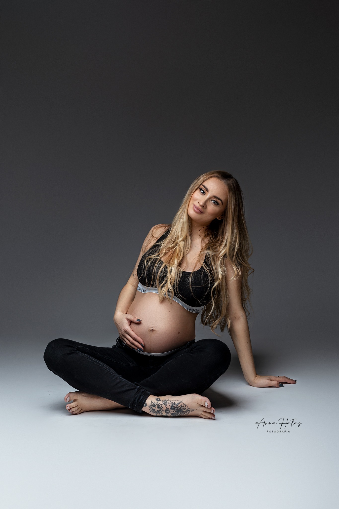 Kobieta w ciąży, siedząca w pozycji lotosu na szarym tle, ubrana w czarny sportowy biustonosz i dżinsy, z tatuażem na stopie, delikatnie dotyka brzucha.