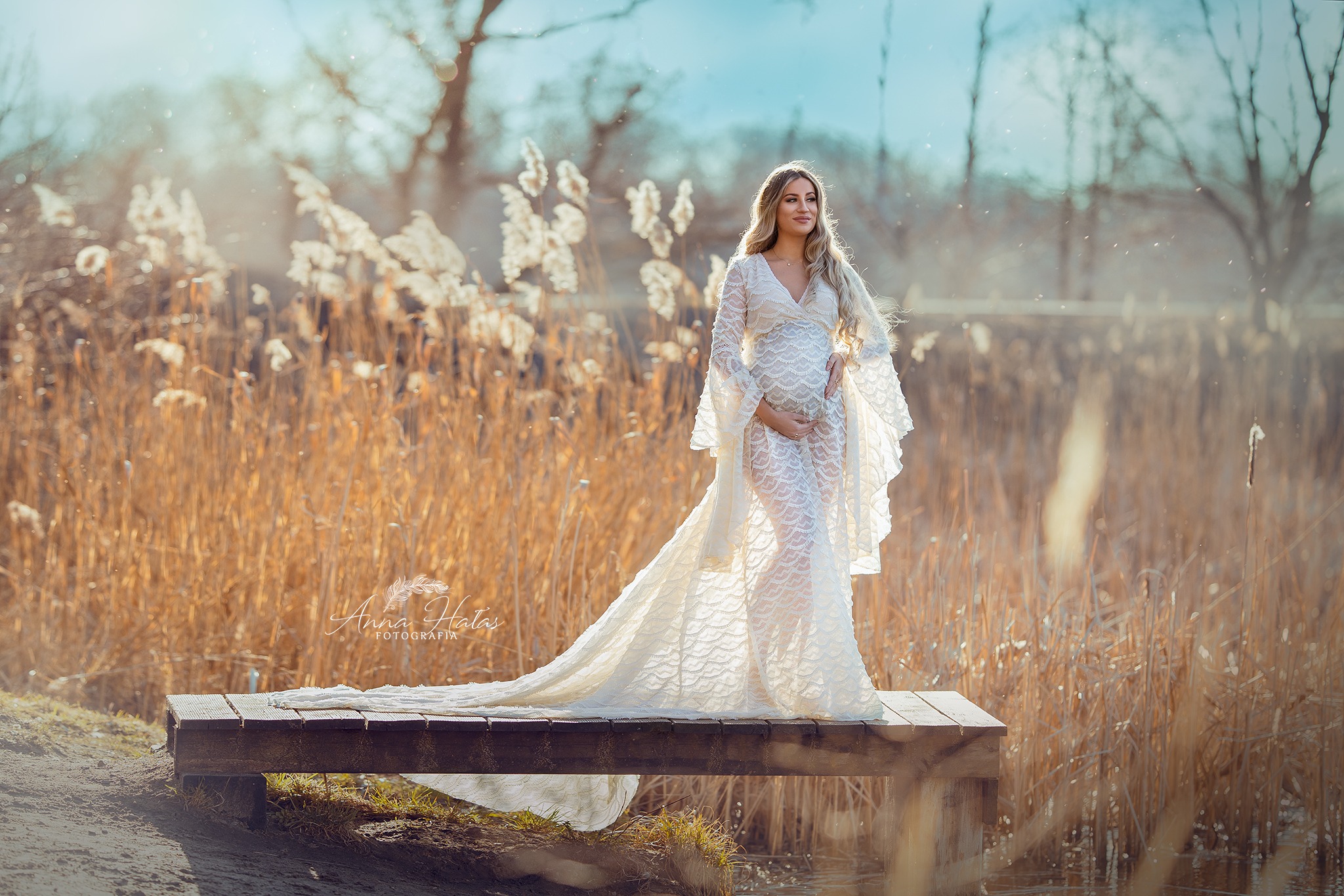 Kobieta w długiej, koronkowej sukni ciążowej, stojąca na drewnianym pomoście na tle trzcin i drzew w złocistym świetle.