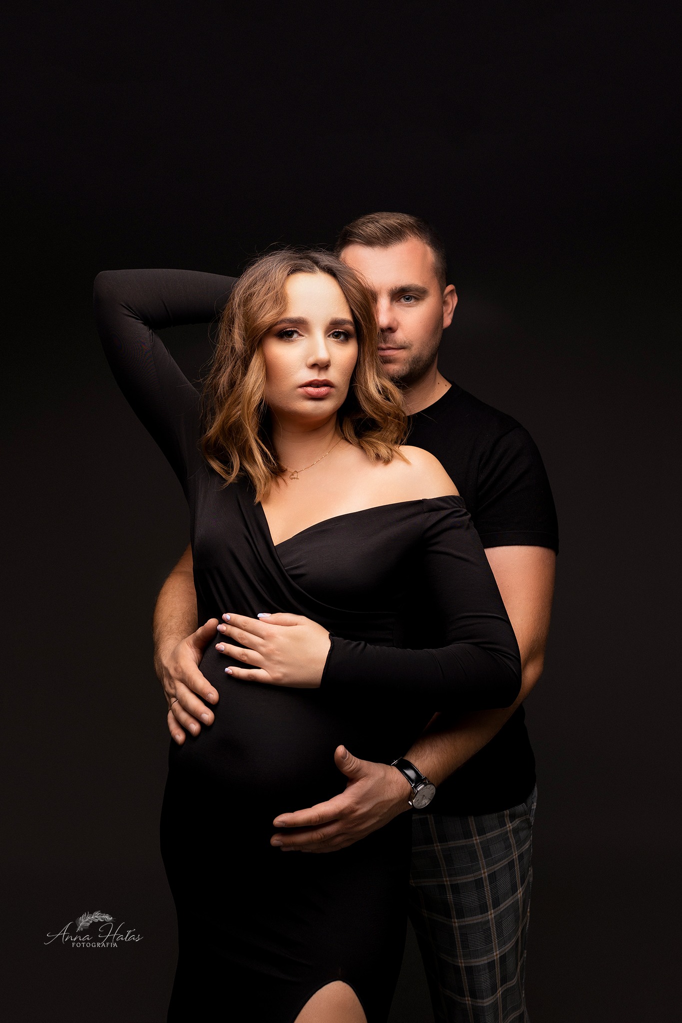 Przyszła mama w czarnej sukience z odkrytym ramieniem, pozuje z partnerem na ciemnym tle, jego ręce obejmują jej brzuch, ona unosi rękę do góry, na szyi ma delikatny złoty łańcuszek.