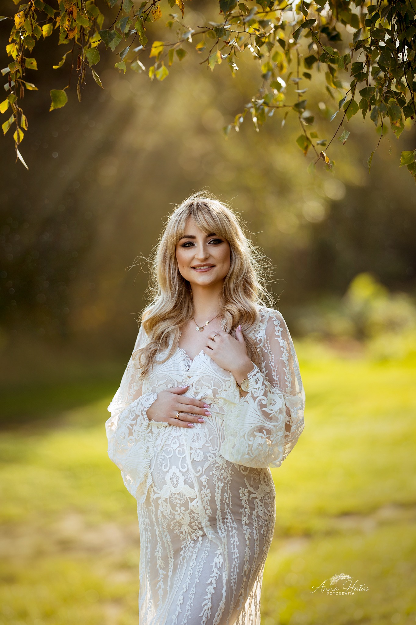Kobieta w ciąży w koronkowej sukni, z rękami na brzuchu, pozuje na tle zieleni, z gałęziami drzew nad głową.