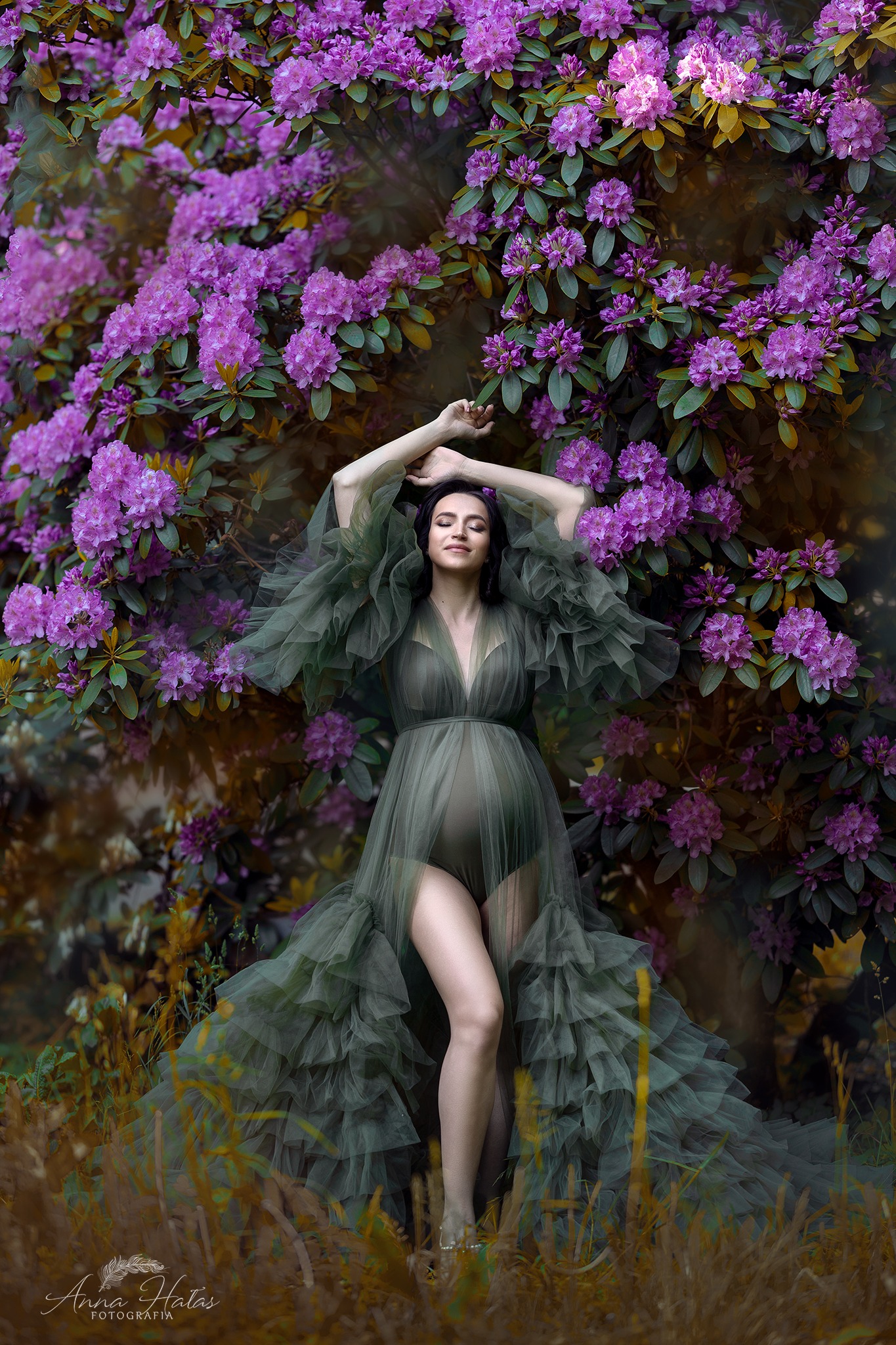 Kobieta w zwiewnej, zielonej sukni pozuje na tle kwitnących, fioletowych rododendronów, ręce uniesione nad głową, delikatny uśmiech na twarzy, widoczny ciążowy brzuszek.