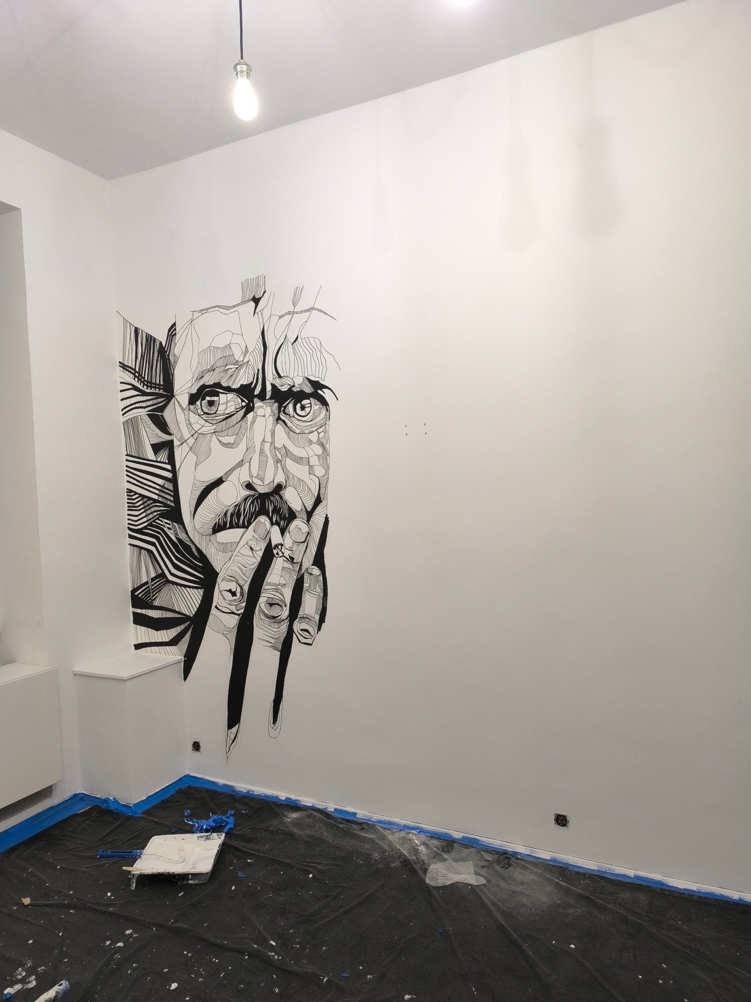 Wnętrze pokoju z białą ścianą ozdobioną czarno-białym, abstrakcyjnym rysunkiem portretowym. Na podłodze folia ochronna z resztkami farby i narzędziami malarskimi.