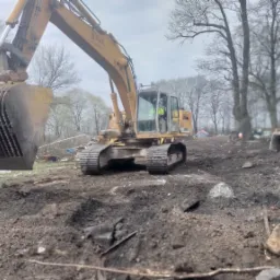 Żółta koparka Liebherr 944 Litronic z ażurową łyżką, pracująca na nierównym, błotnistym terenie z resztkami gruzu i drzewami w tle.