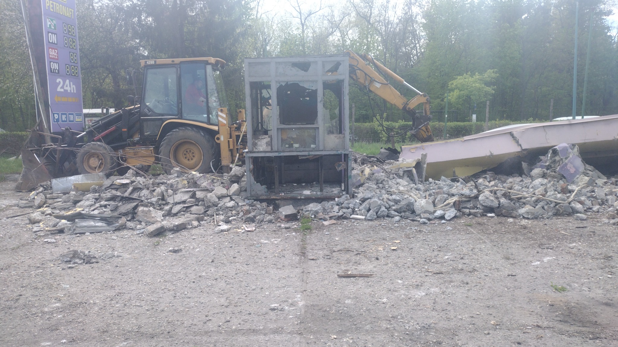 Żółta koparko-ładowarka usuwa gruz i pozostałości po wyburzonym budynku stacji benzynowej z dużym pylonem reklamowym w tle.