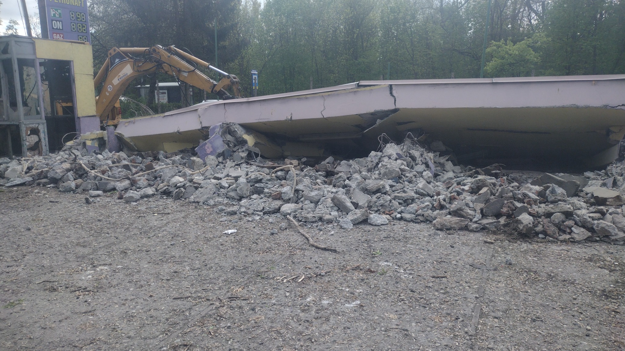 Żółta koparka przy częściowo zburzonym budynku stacji benzynowej, gruz na ziemi, widoczny fragment cennika paliw