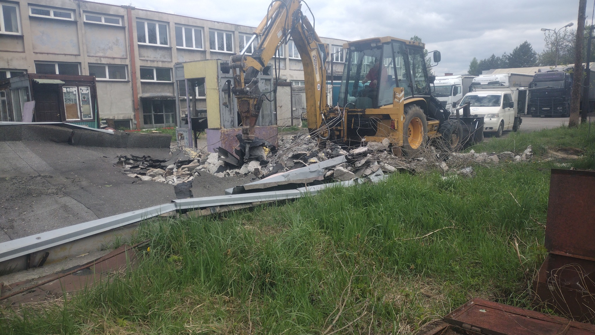 Żółta koparko-ładowarka podczas kruszenia fragmentu budynku, z gruzem na ziemi i budynkiem w tle, widoczne ciężarówki w oddali.