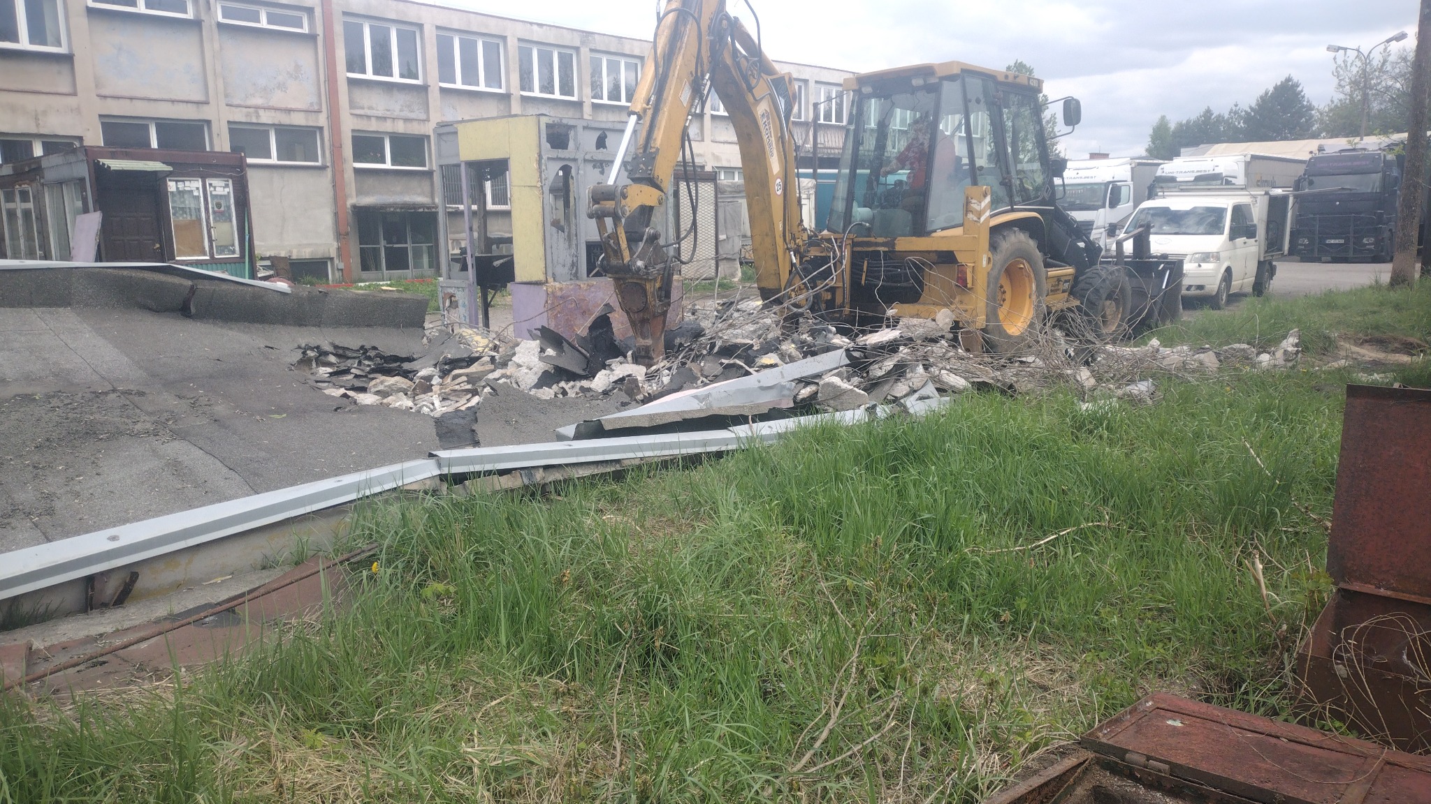 Żółta koparko-ładowarka z hydraulicznym młotem kruszy pozostałości budynku na placu budowy, w tle budynek i zaparkowane samochody dostawcze.