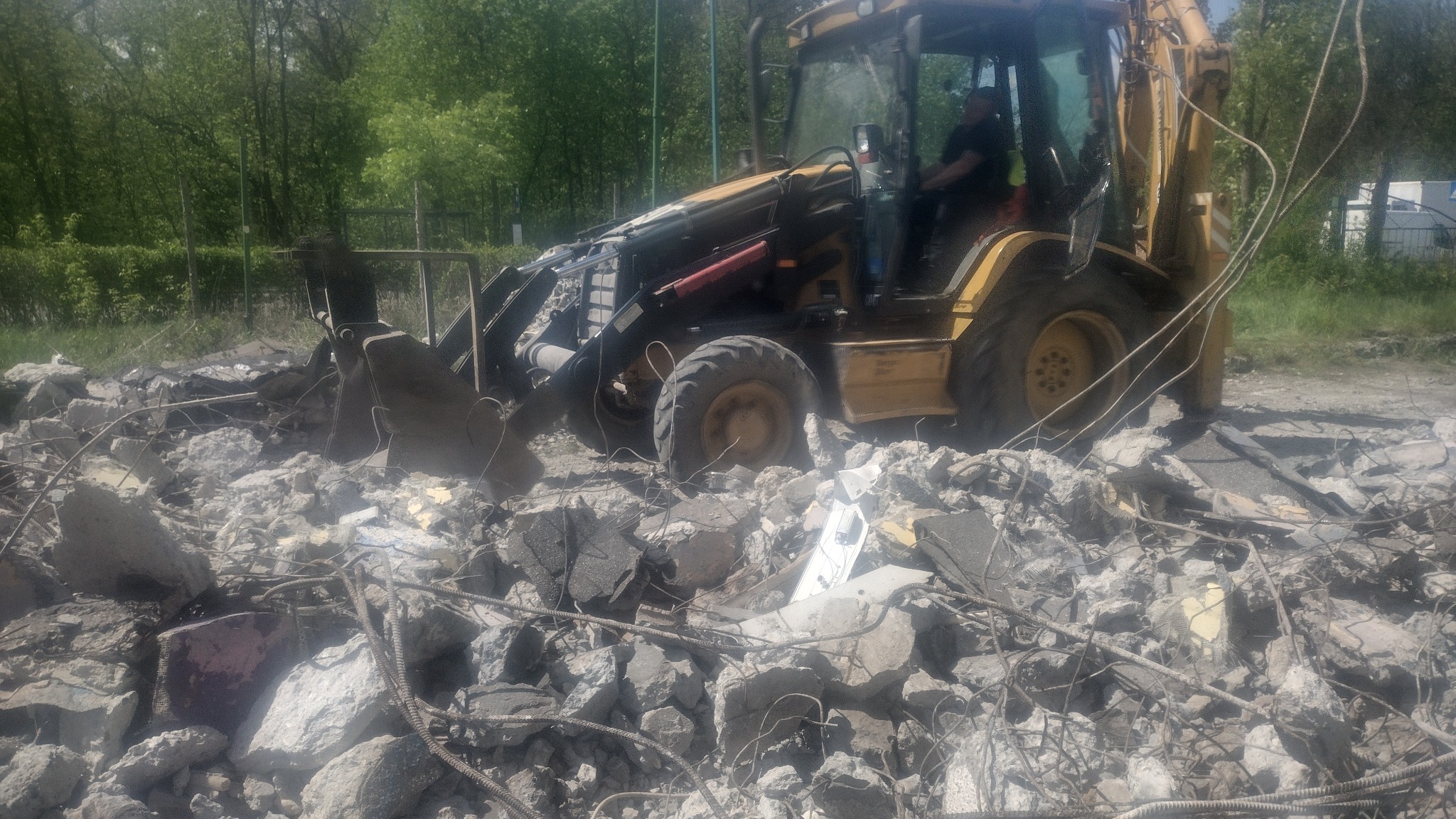 Żółta koparko-ładowarka na stercie gruzu z wystającymi prętami zbrojeniowymi, w tle zieleń drzew.