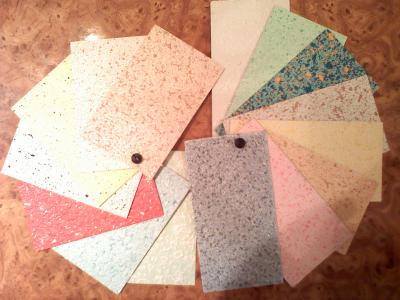 Paleta próbek różnokolorowych materiałów wykończeniowych z widoczną fakturą, spiętych metalowym nitem, ułożonych wachlarzowo na drewnianym blacie.