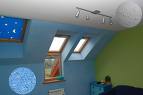 Pokój na poddaszu po remoncie: błękitne ściany, trzy okna dachowe, zielona ściana w rogu, biała lampa sufitowa z czterema reflektorami, widoczny fragment grzejnika.