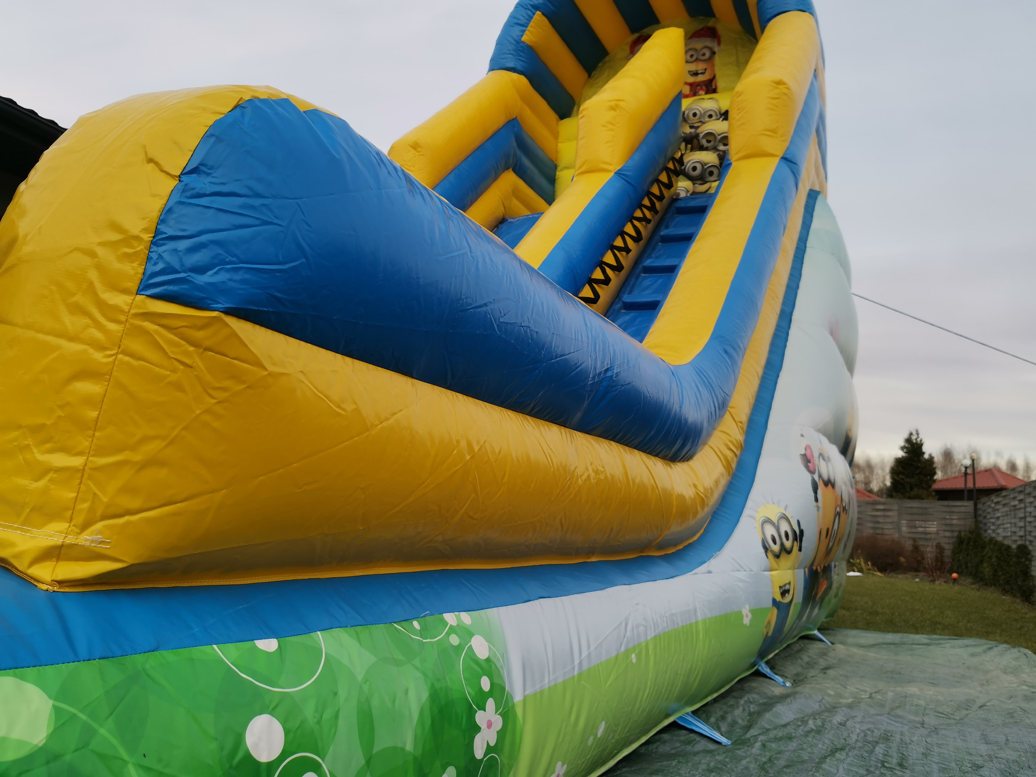 Duża, dmuchana zjeżdżalnia w kolorach żółtym i niebieskim, z motywem Minionków, ustawiona na trawniku przed domem.