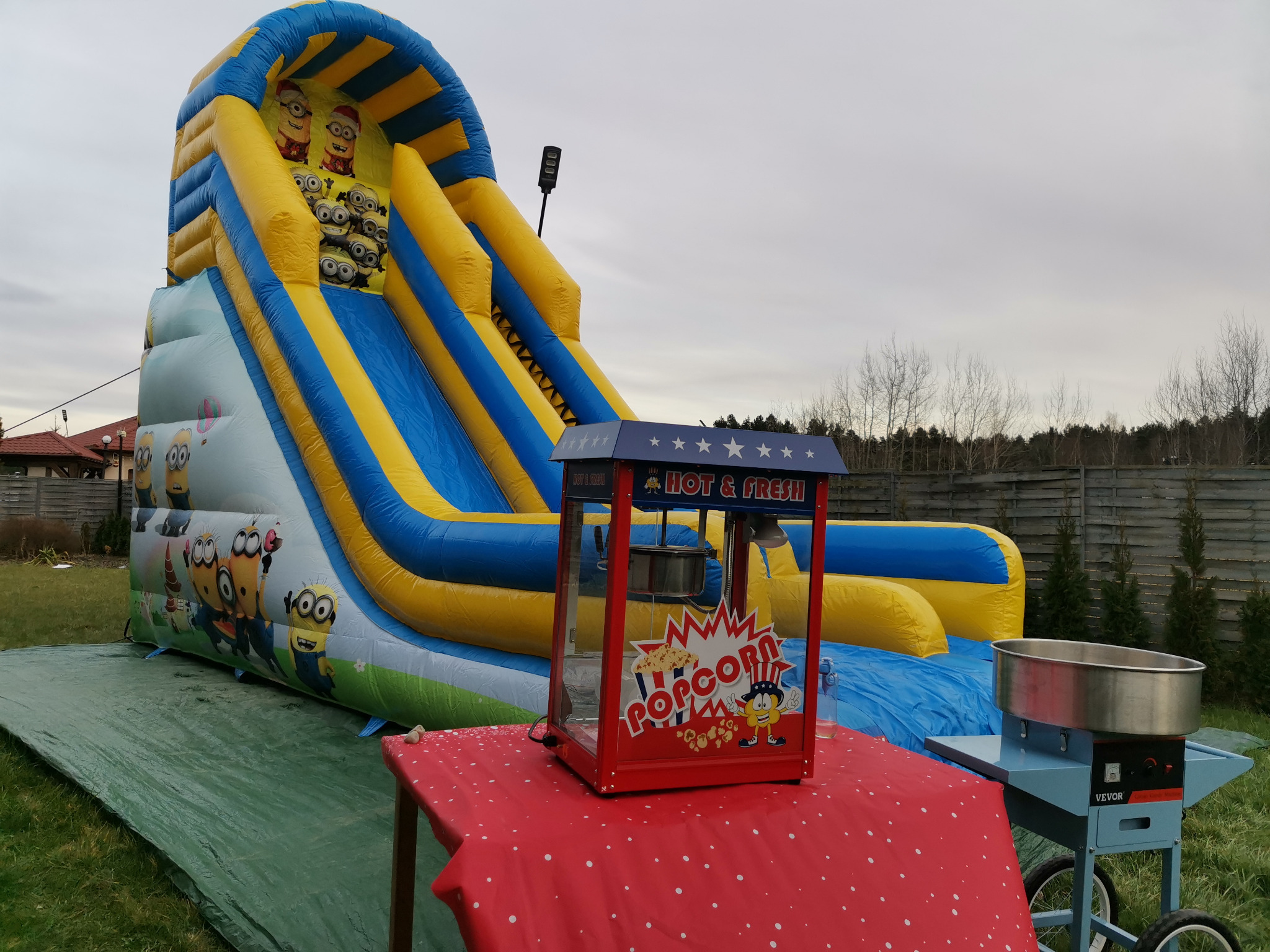 Duży dmuchany zamek ze zjeżdżalnią z motywem Minionków, automat do popcornu i maszyna do waty cukrowej na zewnątrz, ustawione na trawie.
