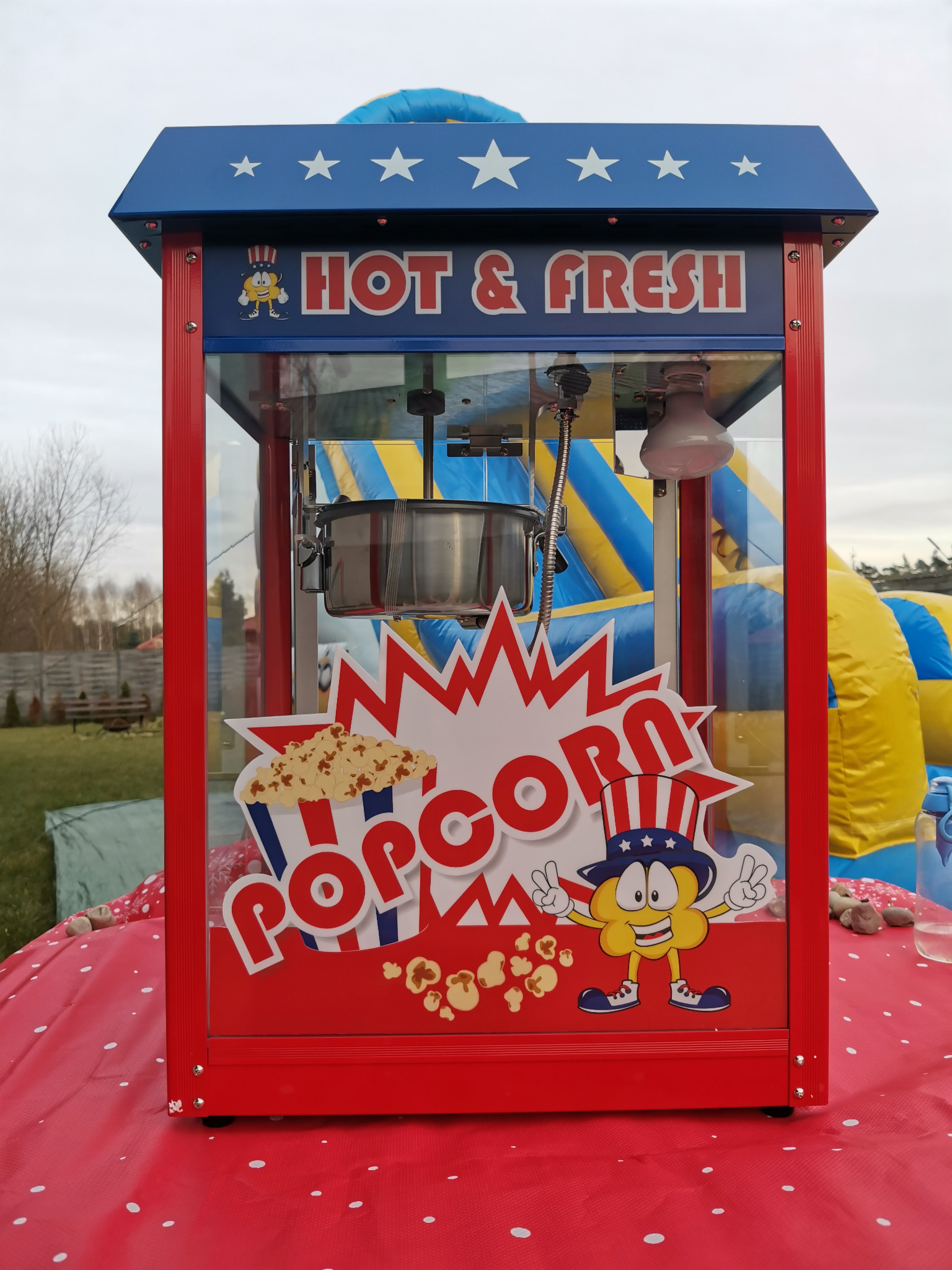 Automat do popcornu z napisem 'Hot & Fresh' i rysunkiem kukurydzy w cylindrze, ustawiony na czerwonym stole w kropki, w tle dmuchany zamek.
