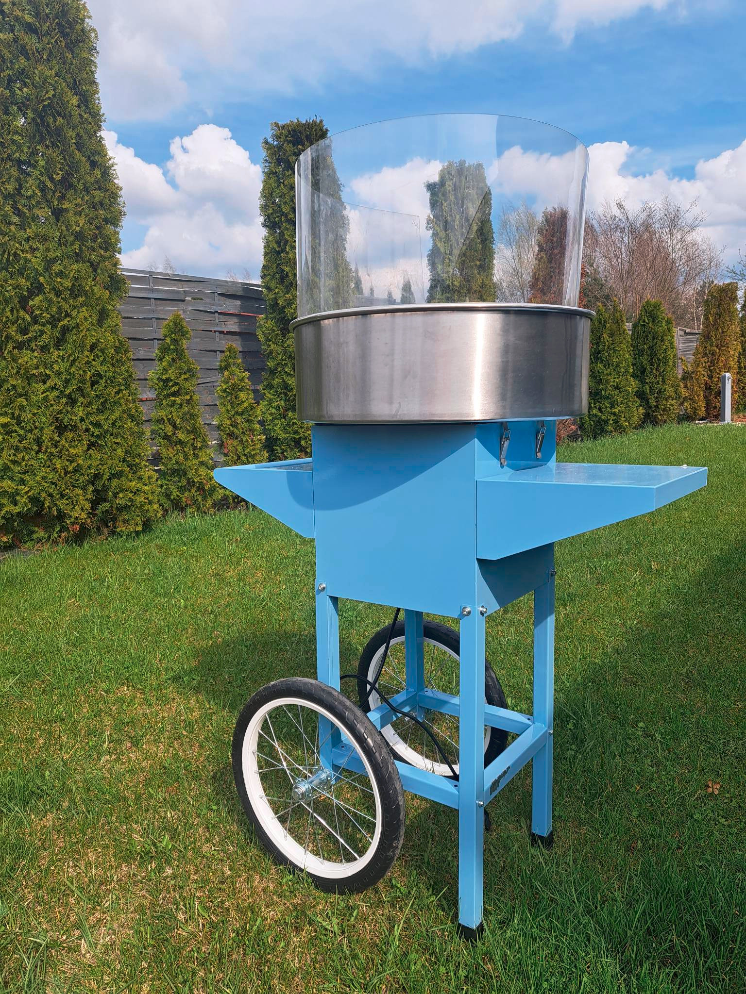 Niebieski wózek do waty cukrowej z przezroczystą osłoną, stojący na trawie w ogrodzie, widoczne koła z białymi oponami.