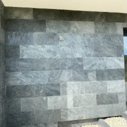 Szara, kamienna elewacja budynku ułożona z prostokątnych płyt, widoczny fragment okna i mały fragment chodnika z białym żwirem.