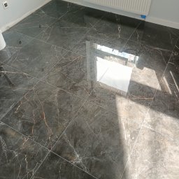 ASBud - Błyszcząca, ciemna podłoga z płytek imitujących marmur z wyraźnymi żyłami w odcieniach bieli i brązu, świeżo po ułożeniu, z widocznym grzejnikiem na ścianie i odbiciem światła słonecznego.
