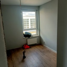 ASBud - Pokój po remoncie z ułożonymi panelami podłogowymi, widoczna torba narzędziowa, włącznik światła i wystające kable z sufitu.