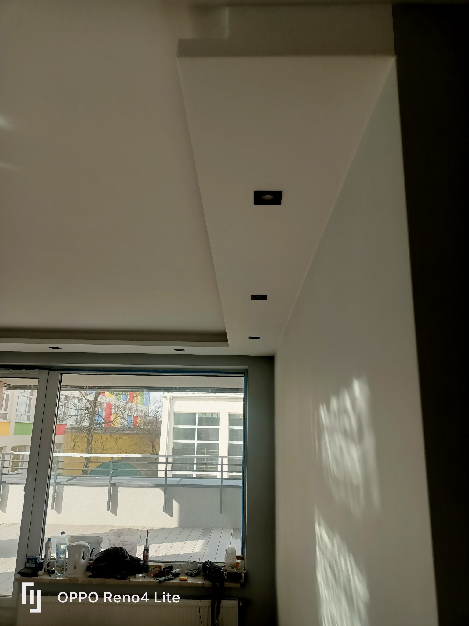 Nowoczesne wykończenie sufitu podwieszanego z trzema czarnymi reflektorami punktowymi i gładkimi ścianami, widoczne narzędzia malarskie na parapecie okna.