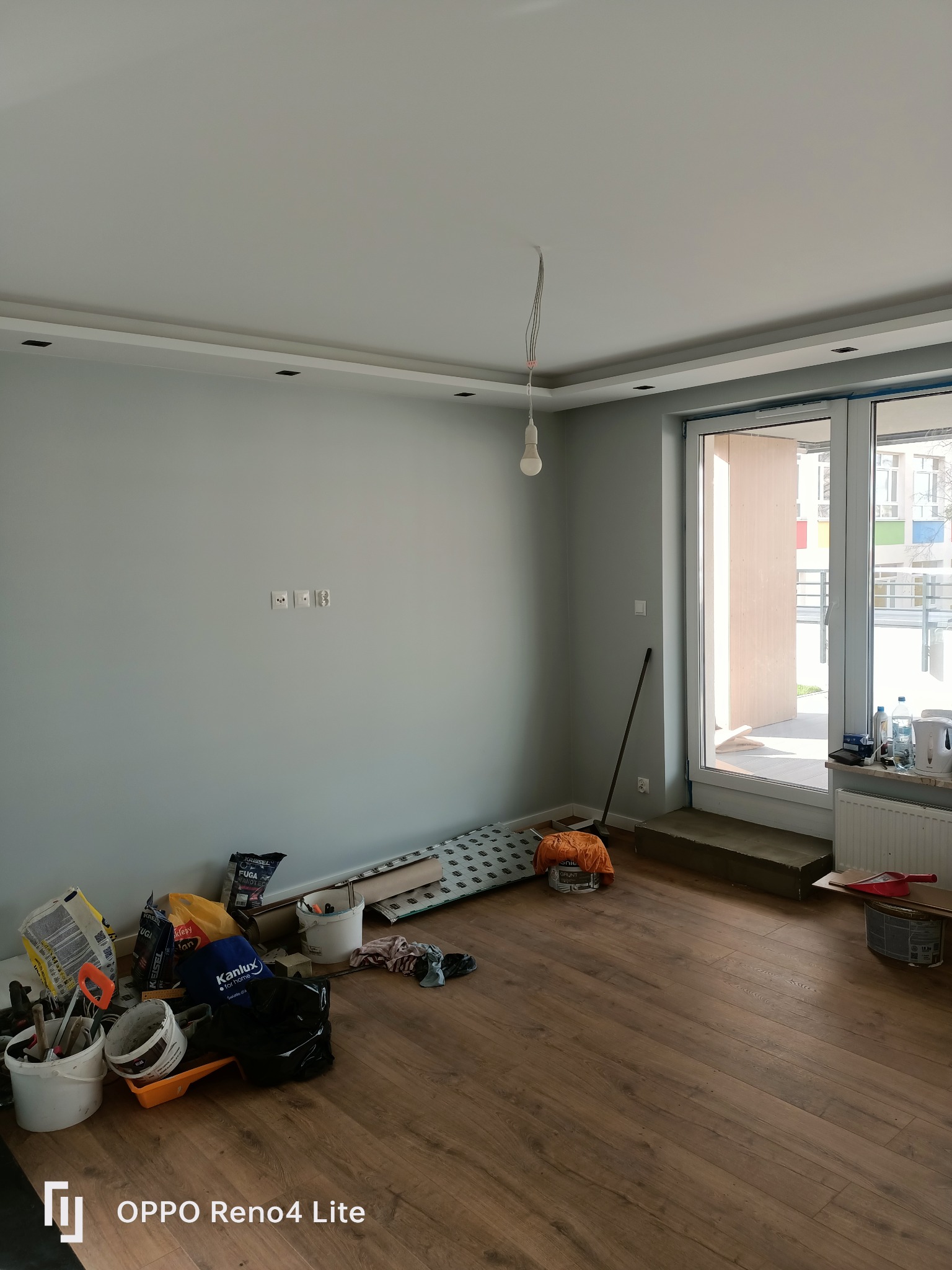 Pokój w trakcie remontu z widocznymi materiałami budowlanymi, workami po klejach, narzędziami, częściowo położoną podłogą z paneli i surowym oświetleniem wiszącym na kablu.