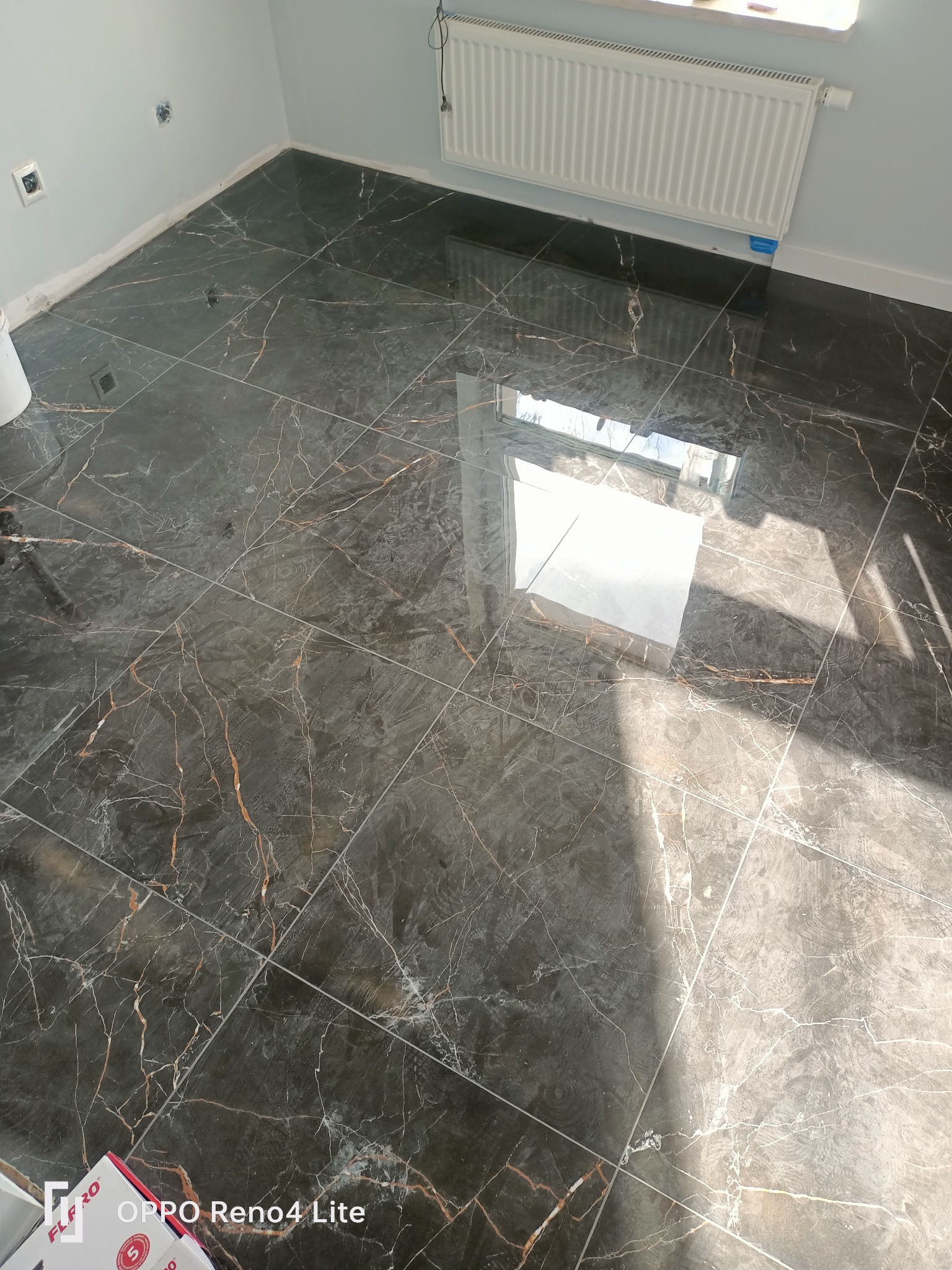 Błyszcząca, ciemna podłoga z płytek imitujących marmur z wyraźnymi żyłami w odcieniach bieli i brązu, świeżo po ułożeniu, z widocznym grzejnikiem na ścianie i odbiciem światła słonecznego.