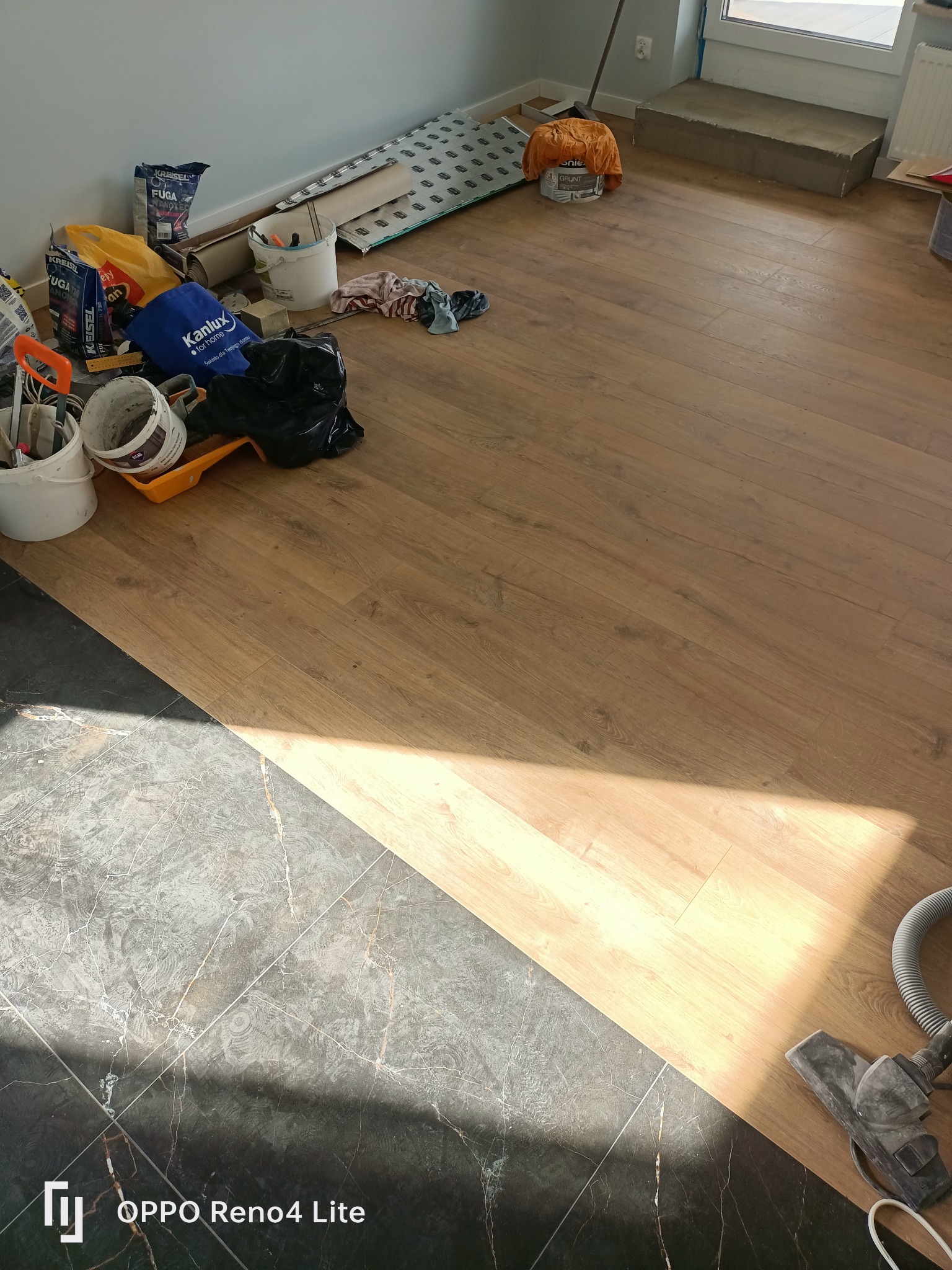 Fragment pokoju podczas prac wykończeniowych: podłoga z paneli przechodząca w ciemne płytki, obok narzędzia, wiadro z zaprawą, worki z klejem i fuga, oraz odkurzacz.