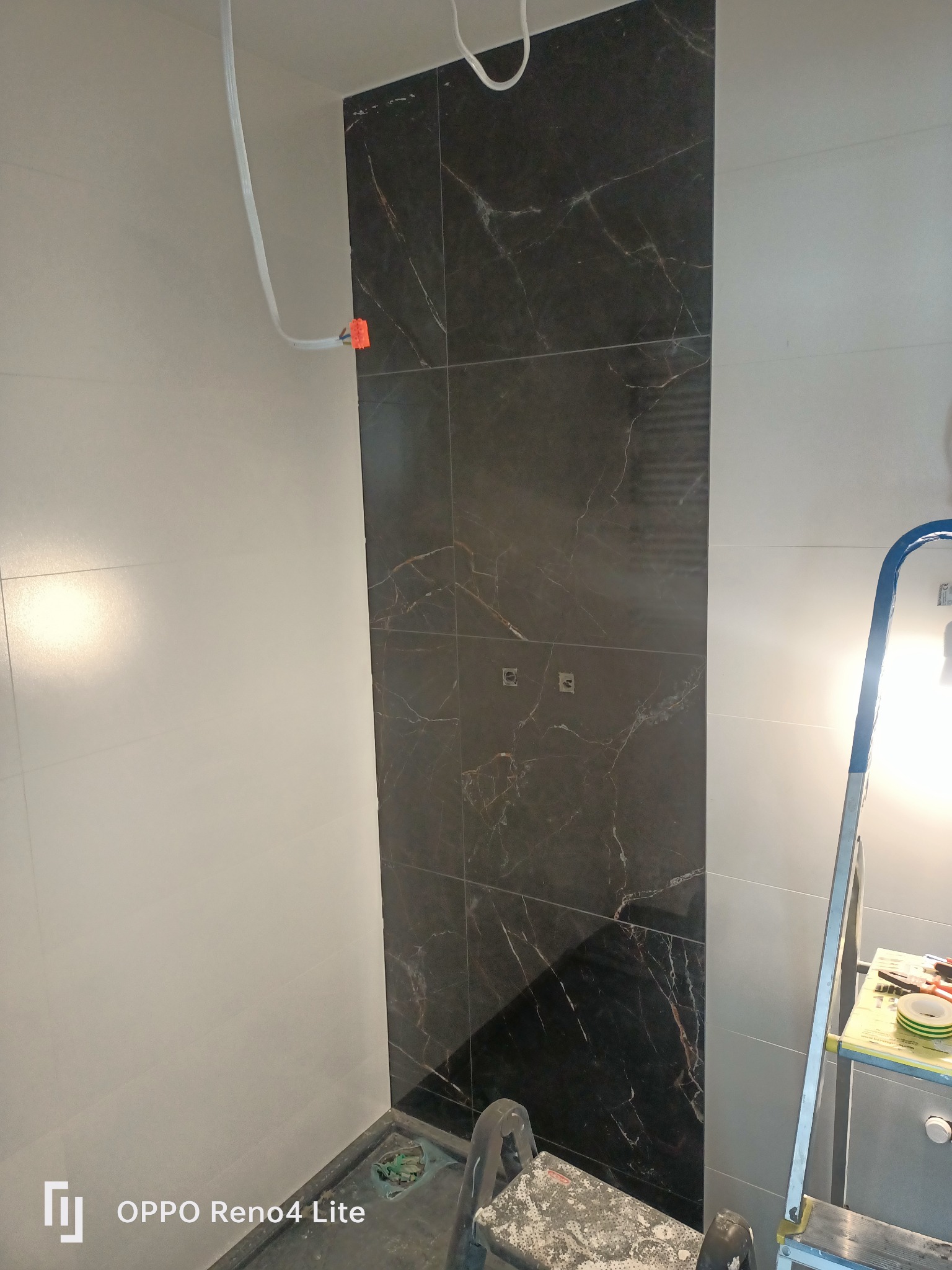 Częściowo wykończona ściana w łazience z czarnymi płytkami imitującymi marmur, widoczna instalacja elektryczna, drabina malarska i narzędzia glazurnicze.