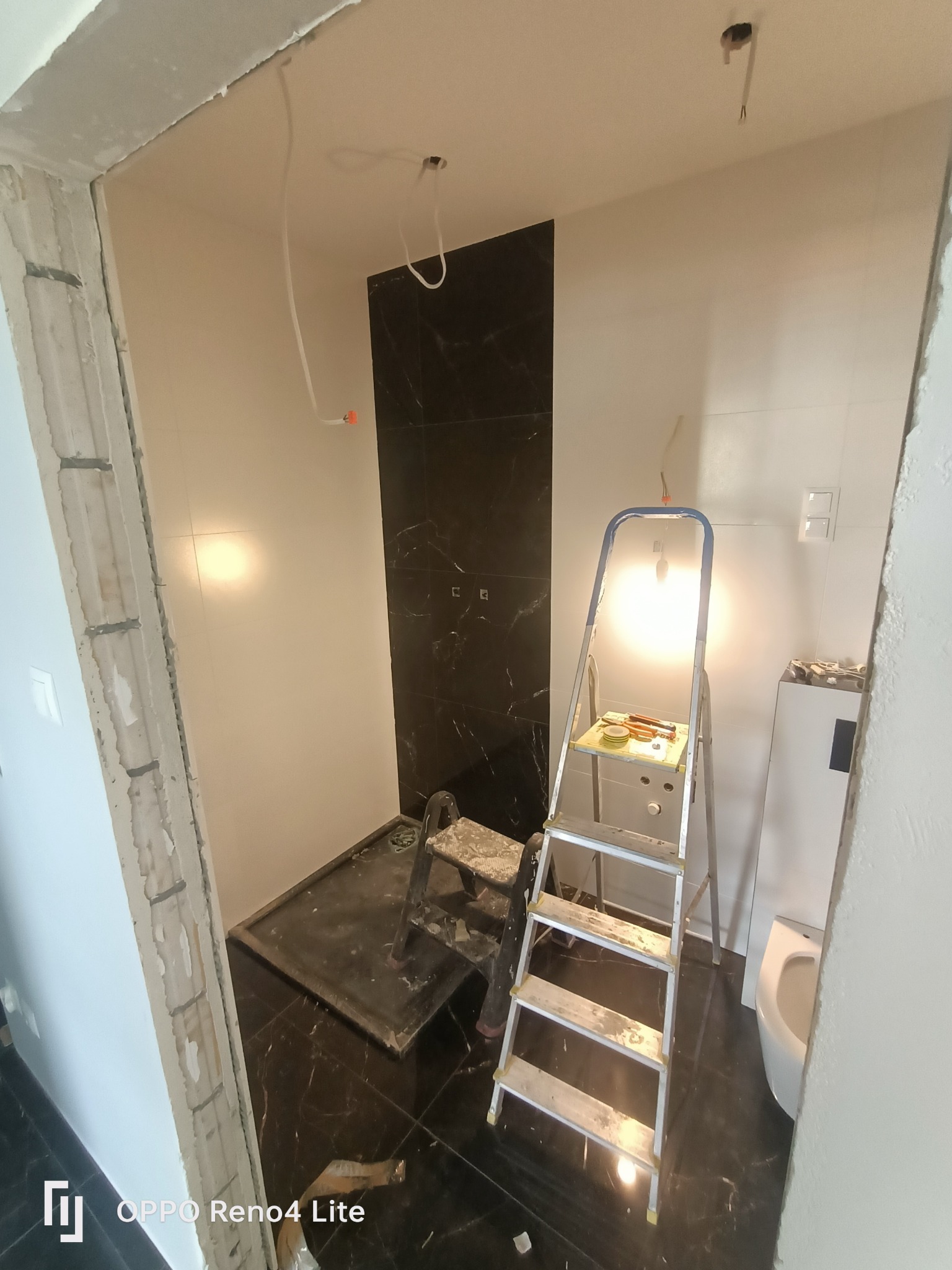 Łazienka w trakcie remontu: czarne marmurowe płytki pod prysznicem i na podłodze, białe ściany, drabina z narzędziami, widoczne przewody elektryczne wystające z sufitu.