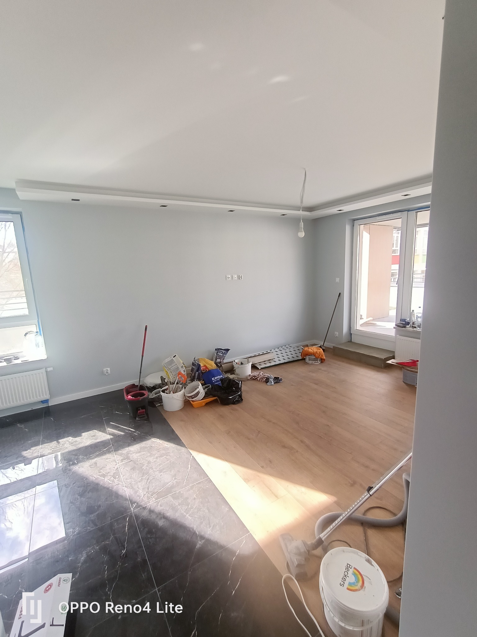 Pokój w trakcie prac wykończeniowych: pomalowane ściany na szaro, częściowo ułożone panele podłogowe, widoczne narzędzia malarskie, pędzle, wiadro z farbą Beckers i odkurzacz.