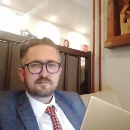 Mężczyzna w marynarce w kratę i krawacie, z laptopem na kolanach, siedzący w kawiarni z obrazami na ścianie w tle.