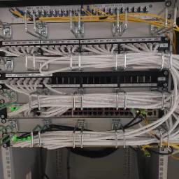 Szczegółowe ujęcie szafy rack z patch panelem i switchami sieciowymi, uporządkowane kable światłowodowe i UTP w metalowych uchwytach, diody LED sygnalizujące pracę urządzeń.