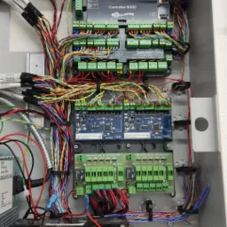 Panel sterowania systemem alarmowym Gallagher Controller 6000 z licznymi przewodami, modułami rozszerzeń i akumulatorem YUCEL, zainstalowany w szarej szafce.
