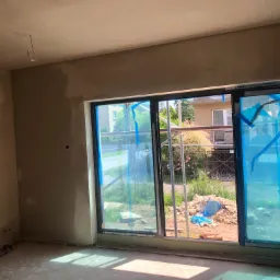 Wnętrze pomieszczenia w trakcie prac wykończeniowych, ściany pokryte surowym tynkiem, okna zabezpieczone folią ochronną, widoczne przewody elektryczne wystające z sufitu.