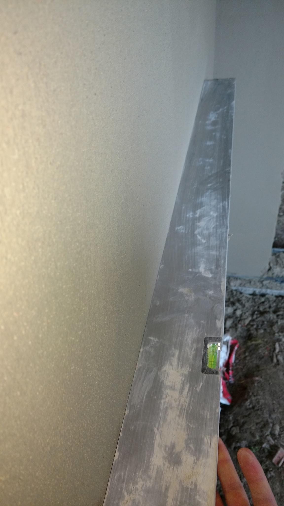 Świeżo nałożona, jasna wyprawa tynkarska na ścianie, sprawdzana poziomicą aluminiową z widocznym pęcherzykiem powietrza, fragment dłoni w dolnym rogu.