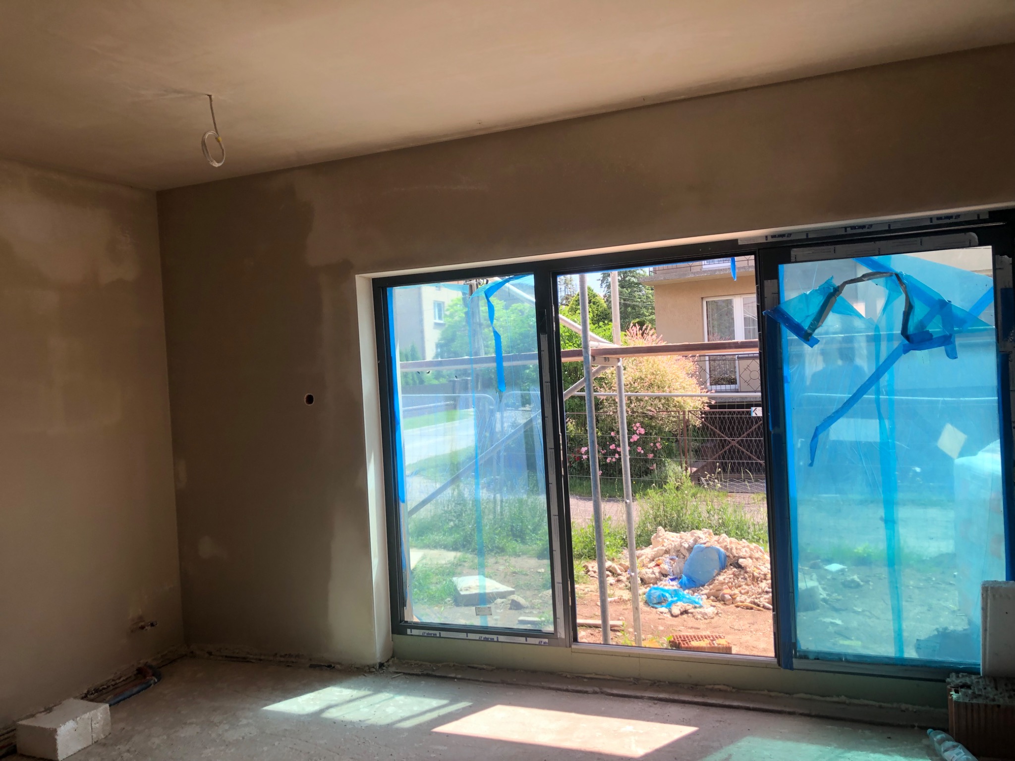 Wnętrze pomieszczenia w trakcie prac wykończeniowych, ściany pokryte surowym tynkiem, okna zabezpieczone folią ochronną, widoczne przewody elektryczne wystające z sufitu.