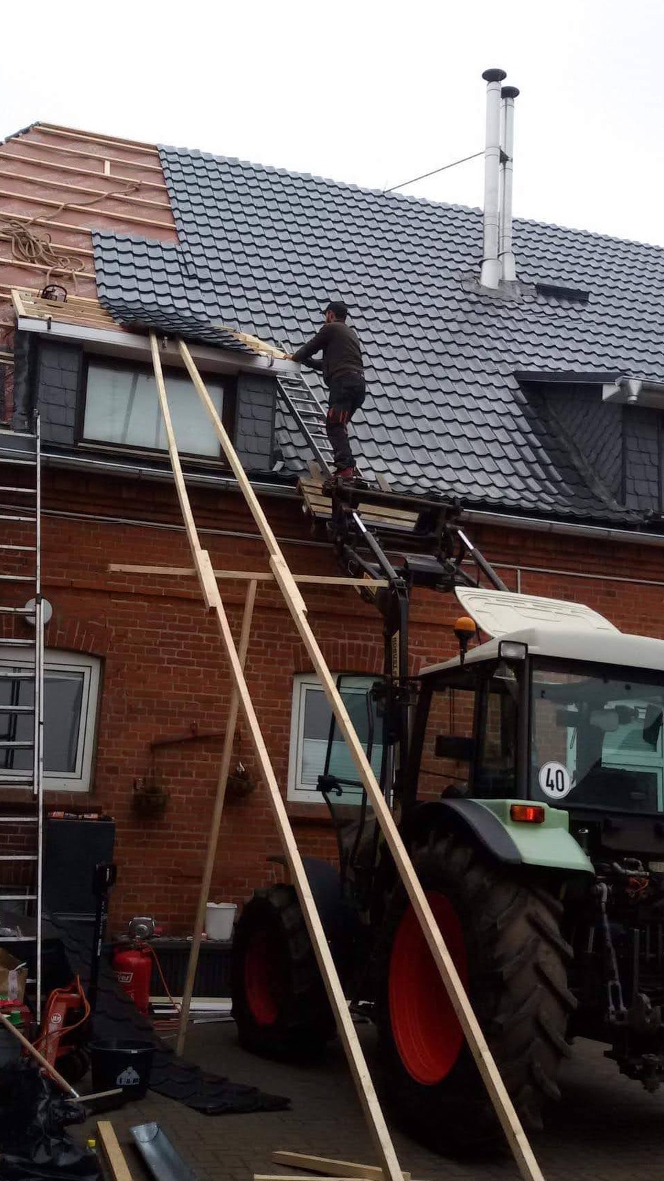 Pracownik na drabinie zamontowanej na traktorze układa dachówkę na częściowo zdjętym dachu budynku z czerwonej cegły; widoczne drewniane rusztowania i komin