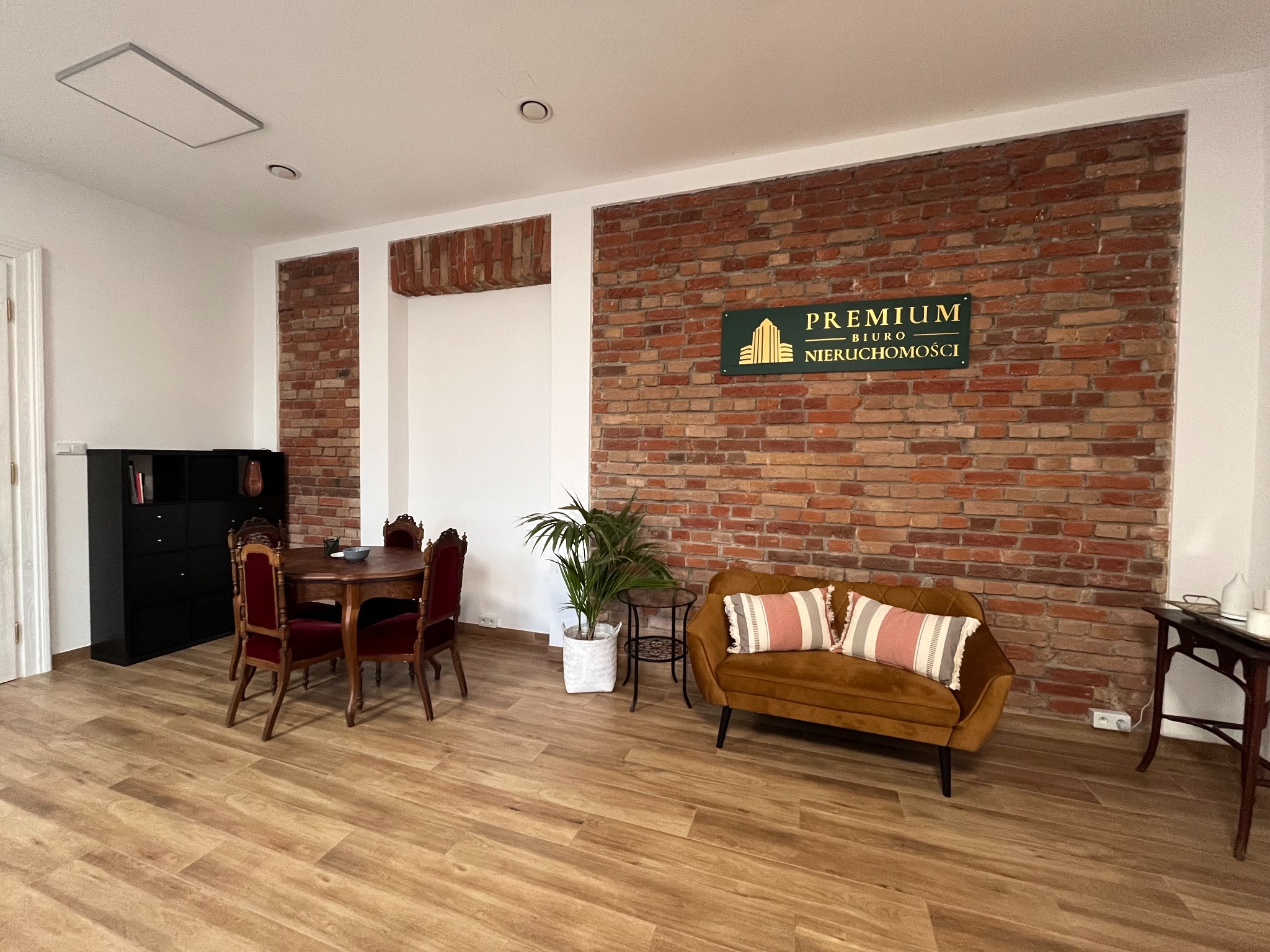 Wnętrze biura nieruchomości z ceglaną ścianą, logo firmy Premium Biuro Nieruchomości, drewnianym stołem i krzesłami oraz welurową kanapą z poduszkami w paski.