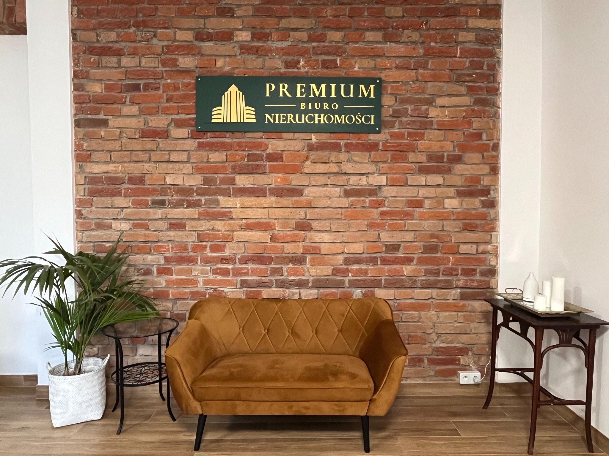 Wnętrze biura nieruchomości z ceglaną ścianą, sofą w kolorze musztardowym, stolikiem z dekoracjami i rośliną doniczkową; na ścianie zielony szyld z napisem 'PREMIUM BIURO NIERUCHOMOŚCI' w kolorze...