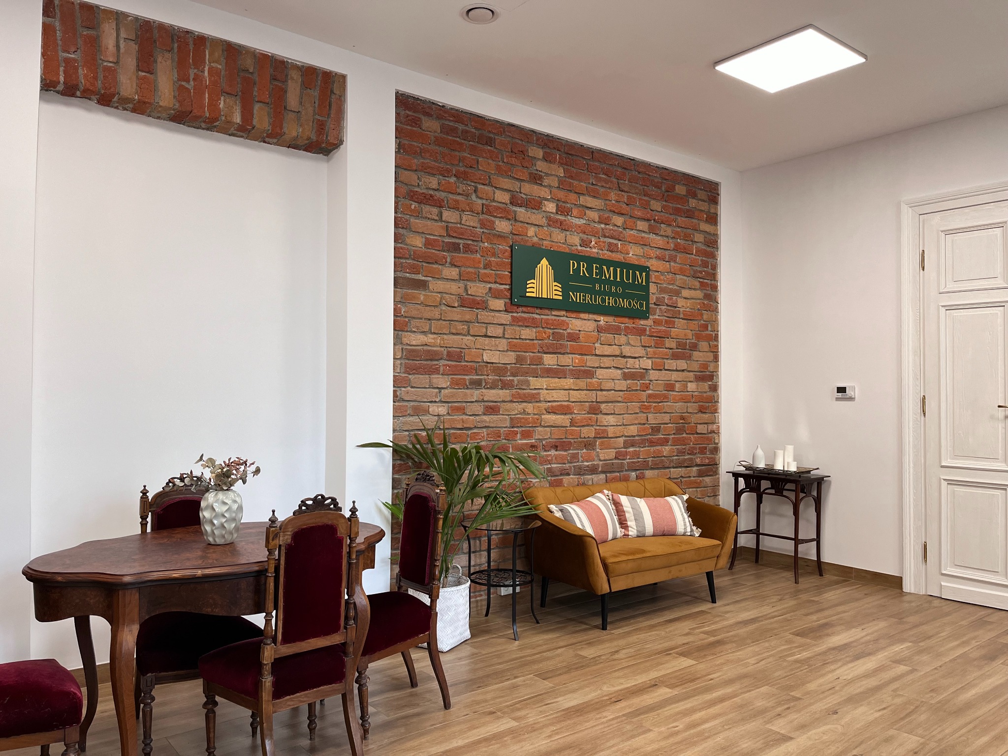 Wnętrze biura nieruchomości: stół z krzesłami w stylu vintage, kanapa w kolorze musztardowym, ściana z cegły z logo firmy 'Premium Biuro Nieruchomości'.