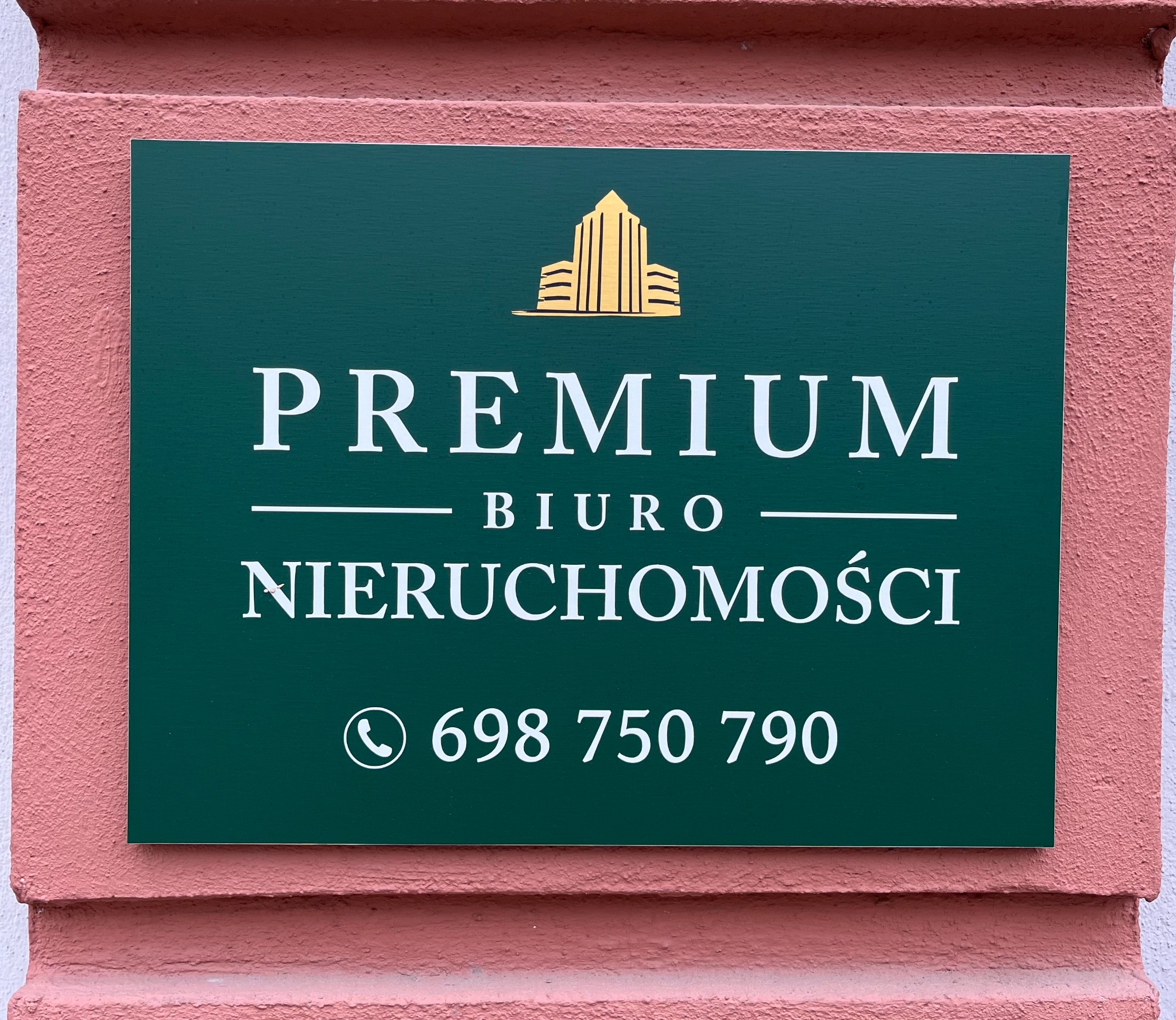 Tablica informacyjna biura nieruchomości PREMIUM w Białymstoku z logo w formie stylizowanego budynku i numerem telefonu na tle różowej ściany.