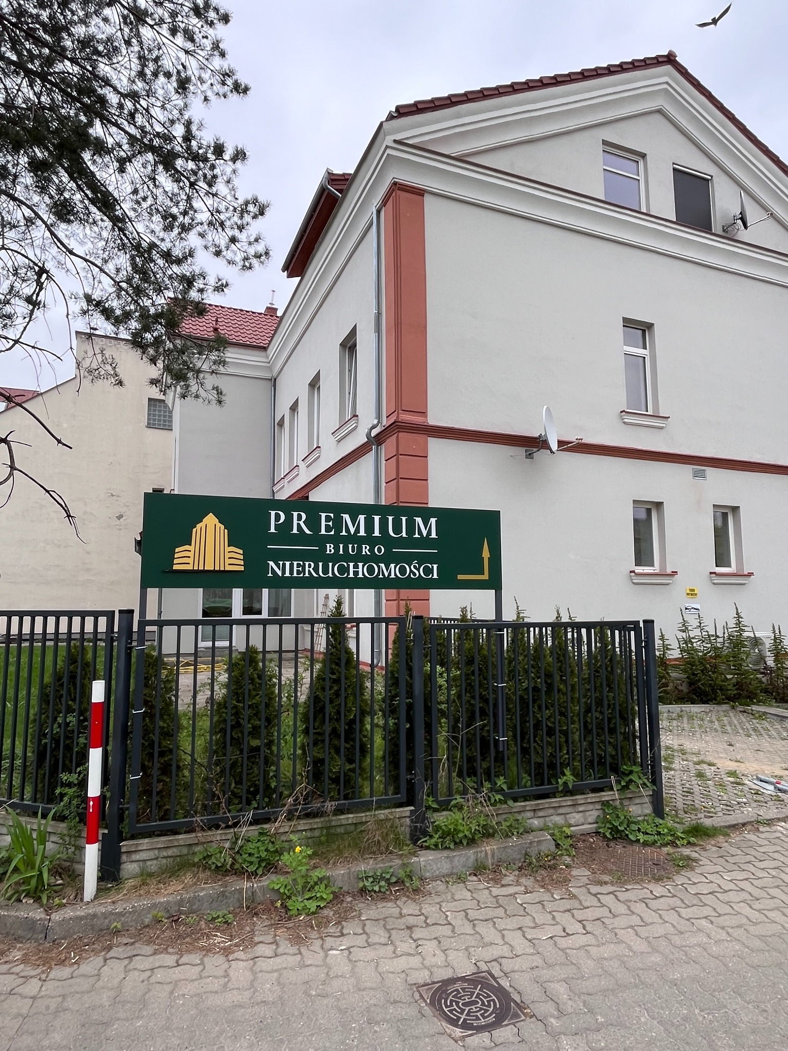 Zewnętrzny widok biura nieruchomości 'Premium Biuro Nieruchomości' z zielonym szyldem z logo złotych budynków, otoczonego czarnym ogrodzeniem z roślinnością i chodnikiem z kostki brukowej.
